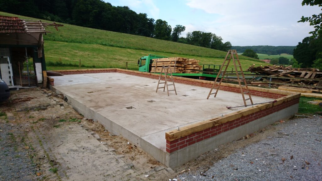 Baustelle mit Betonfundament, Holzleitern und grünem Lastwagen in ländlicher Umgebung. Keine Arbeiter sichtbar.