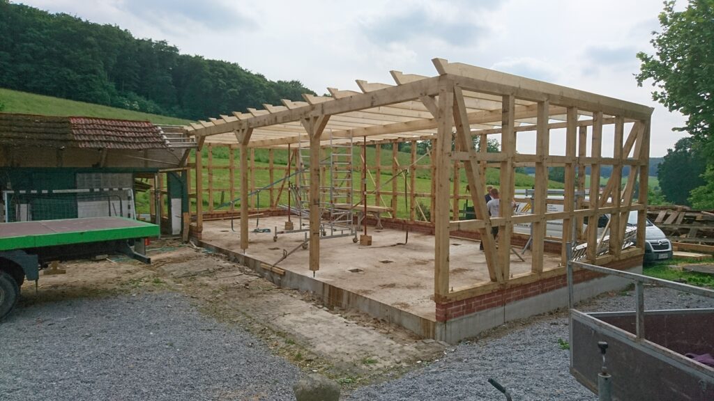 Holzrahmenbau auf ländlicher Baustelle, Arbeiter und Transportfahrzeug, umgeben von grüner Natur.