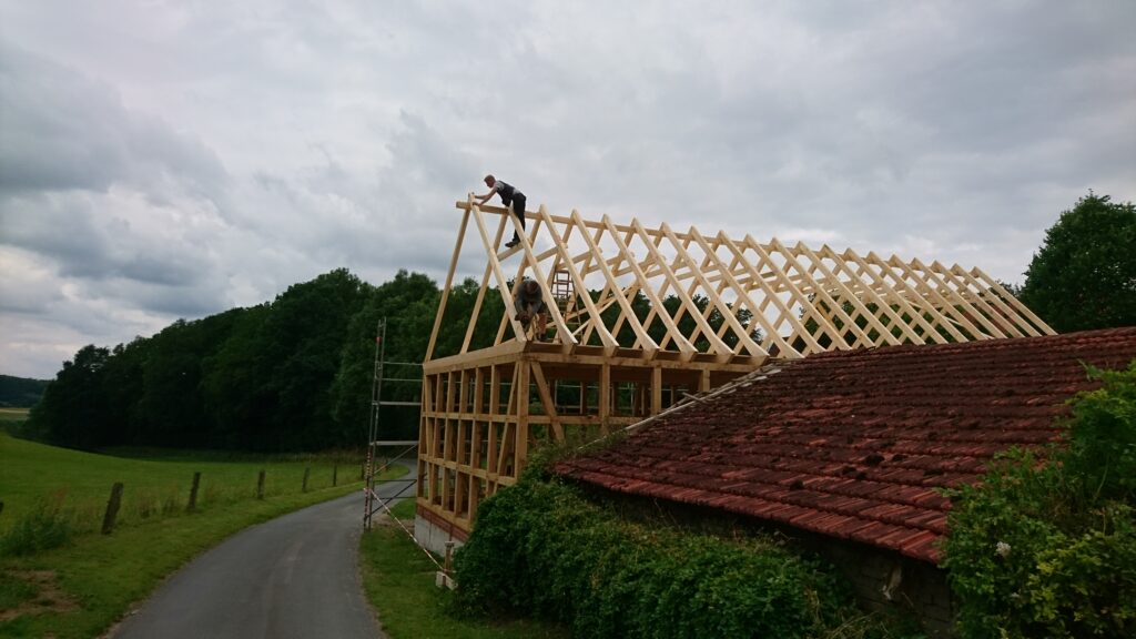 Zwei Personen arbeiten an einem neuen Holzgerüst für ein Dach in ländlicher Umgebung, neben einem alten Gebäude.
