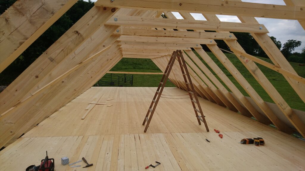 Holzdachkonstruktion im Bau mit Sparren, Leiter und Werkzeugen; ländliche Umgebung im Hintergrund.