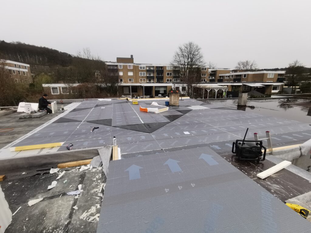 Eine Person arbeitet auf einem Flachdach, verlegt Dämmplatten; umgeben von Werkzeugen und Baumaterialien.
