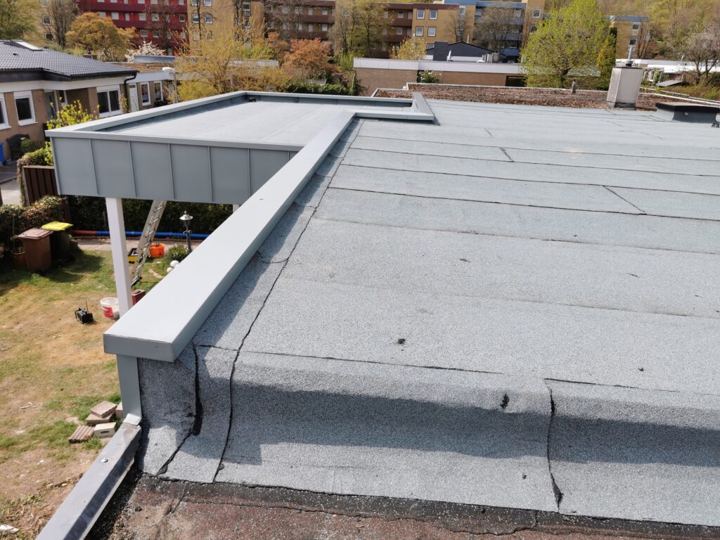 Flachdach mit grauer Dachpappe, umgeben von Bäumen und Gebäuden; ruhiger, sonniger Wohnbereich.