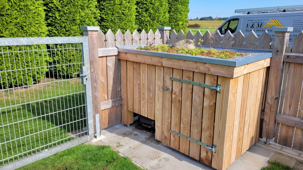 Ein Holzschuppen mit begrüntem Dach neben einem silbernen Metalltor, umgeben von Hecken und Pflastersteinen.