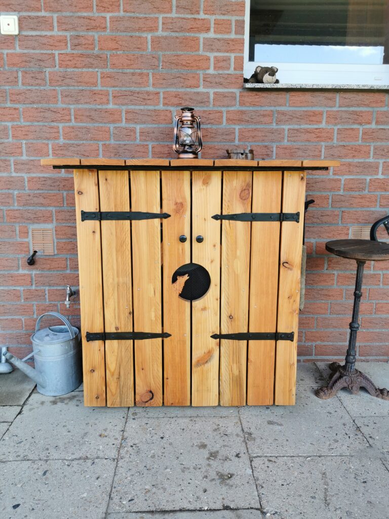 Ein rustikales Holzschränkchen mit Laterne und Gießkanne vor einer roten Backsteinmauer im Freien.