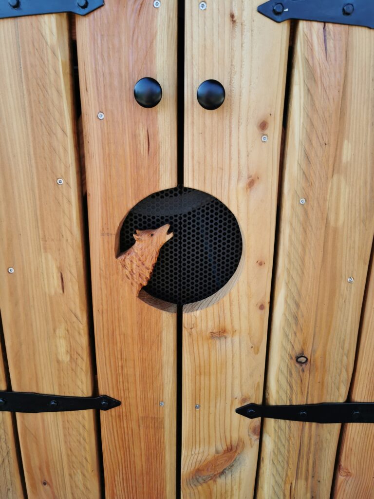 Rustikale Holztür mit schwarzen Griffen und geschnitztem Wolfskopf in einem kreisförmigen Gitter.