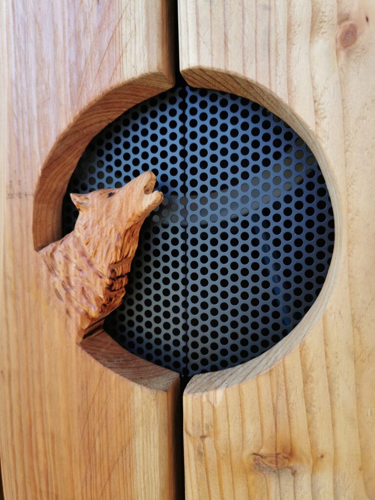 Detaillierte Holzschnitzerei eines heulenden Wolfes in einem Holzrahmen mit schwarzem Metallgitter.