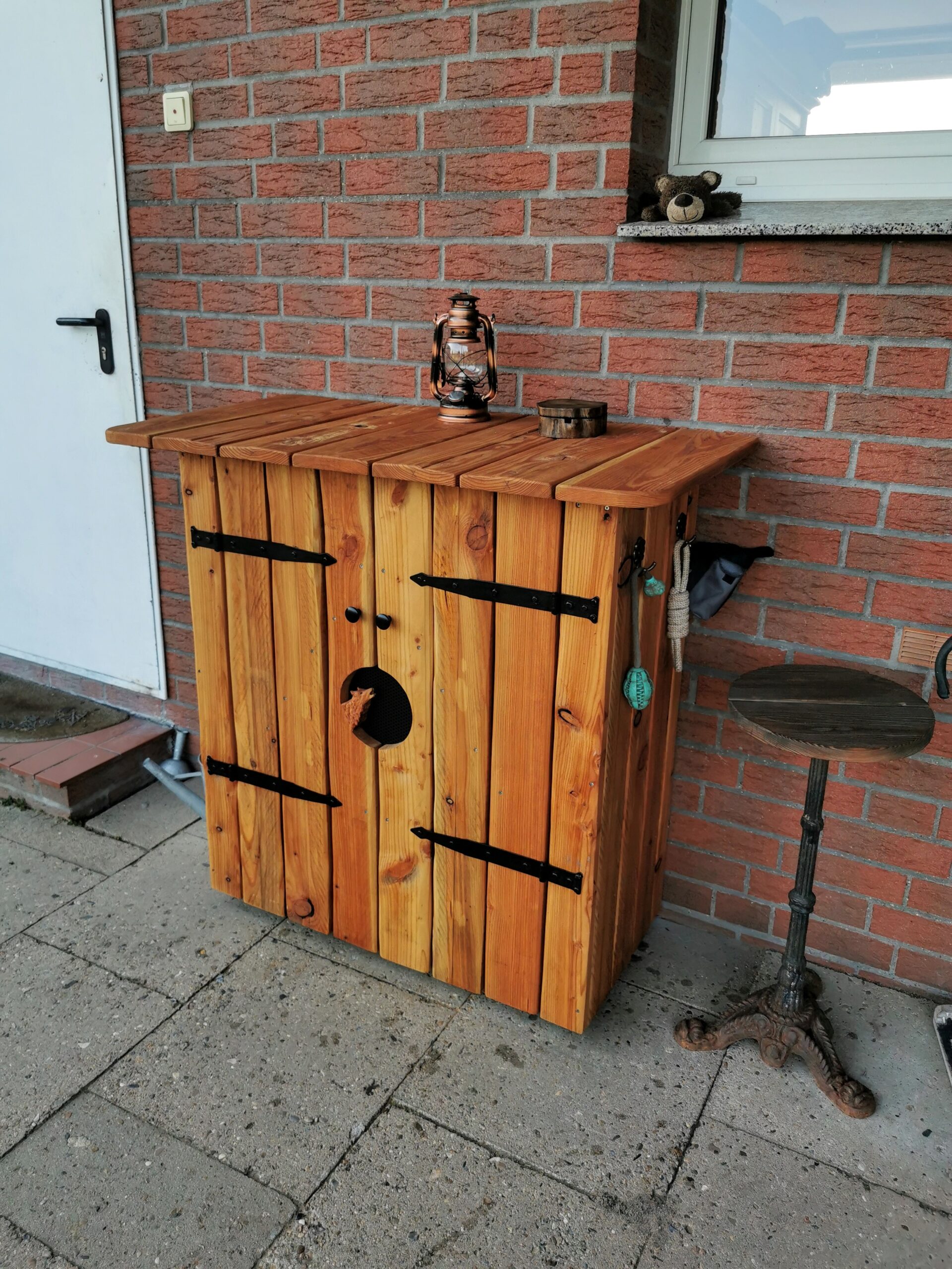 Ein rustikaler Holzschrank mit Laterne, Teddybär und Beistelltisch vor einer roten Backsteinwand.