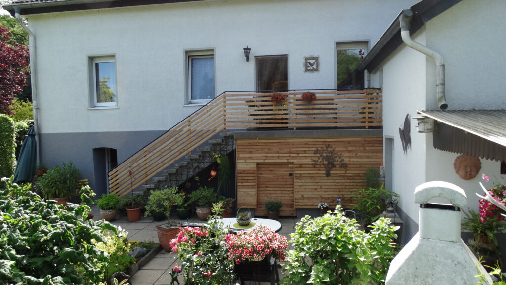 Gepflegter Garten vor einem Wohnhaus mit Balkon, Blumenkästen und einem gemauerten Grill.