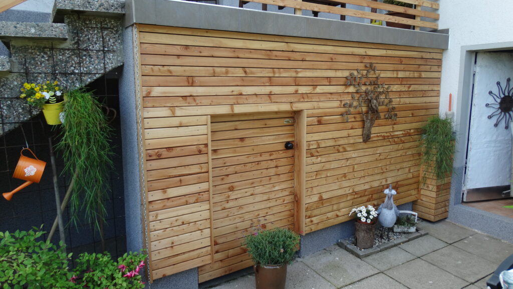 Holzwand mit Tür, Pflanzen, Vogelstatue und orangefarbener Gießkanne; steinerne Treppe links.