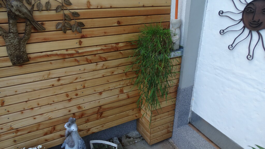 Holzwand mit Metallkunstwerk, Pflanze, Hahnfigur und Sonnenornament in einem ruhigen Außenbereich.