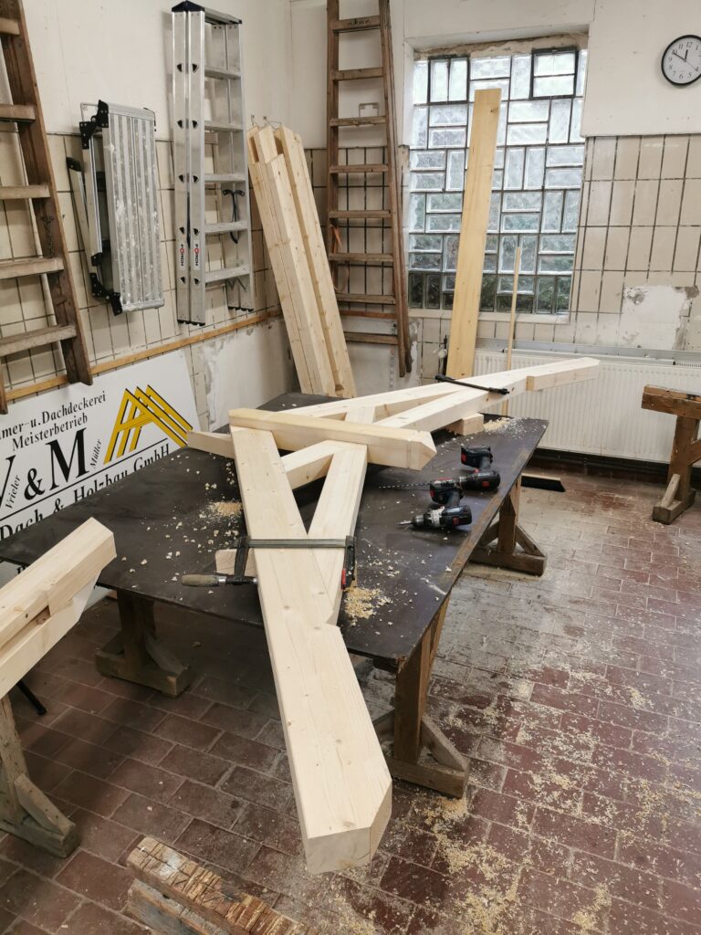Eine Holzwerkstatt mit Holzbalken, Elektroschraubern und Leitern auf einem Arbeitstisch voller Holzspäne.