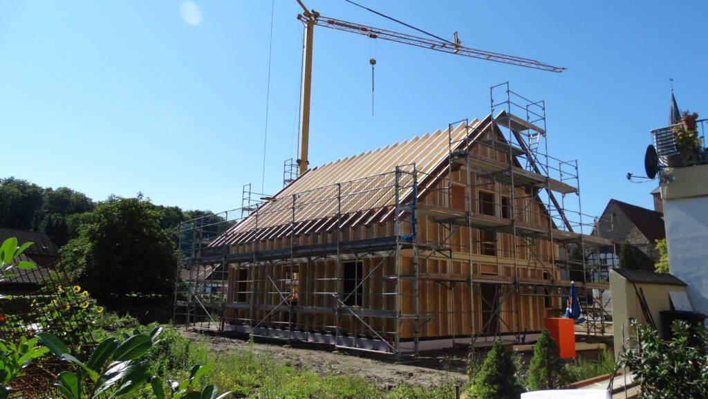 Baustelle mit Holzrahmenhaus, umgeben von Gerüsten und Kran, vor grünem Hintergrund und blauem Himmel.