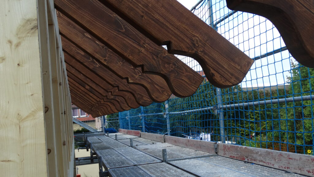 Blick von einer Gerüstplattform auf eine dunkle Holzdachkonstruktion, umgeben von blauen Sicherheitsnetzen.