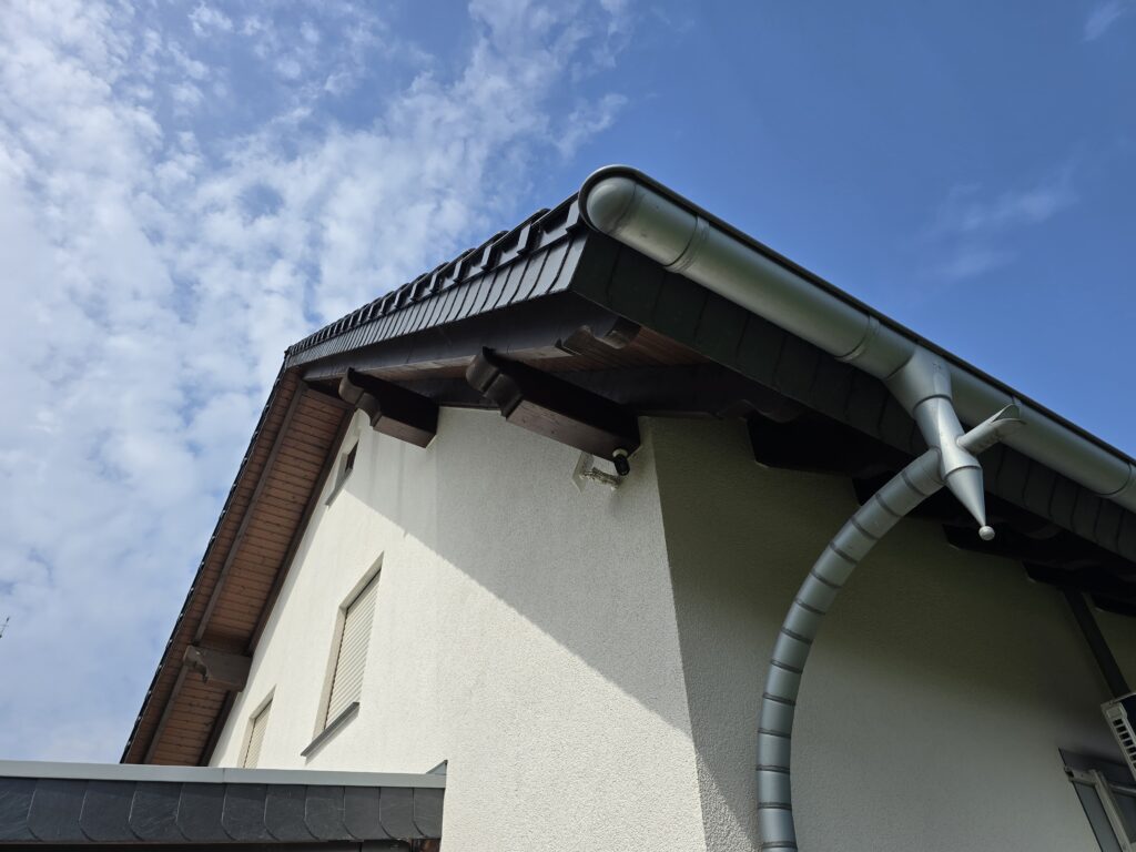Ein Hausdach mit dunklen Ziegeln und silberner Dachrinne vor einem klaren, blauen Himmel.