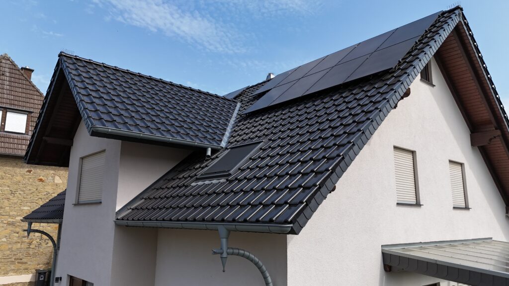 Modernes Haus mit steilem, dunklem Satteldach und Solarpaneelen, umgeben von blauem Himmel.