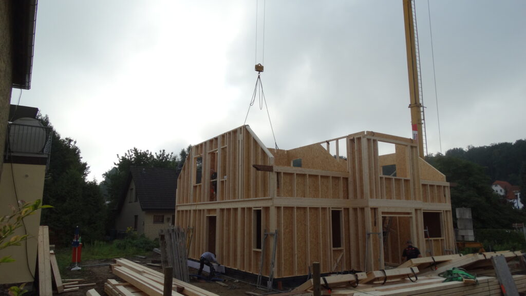 Baustelle eines Holzrahmenhauses mit Kran und Arbeitern unter bewölktem Himmel.