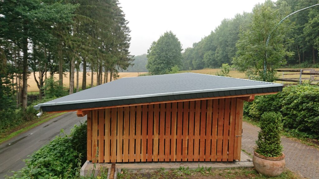 Ein einfaches Holzhäuschen steht ruhig in einer grünen, ländlichen Umgebung mit Bäumen und Büschen.