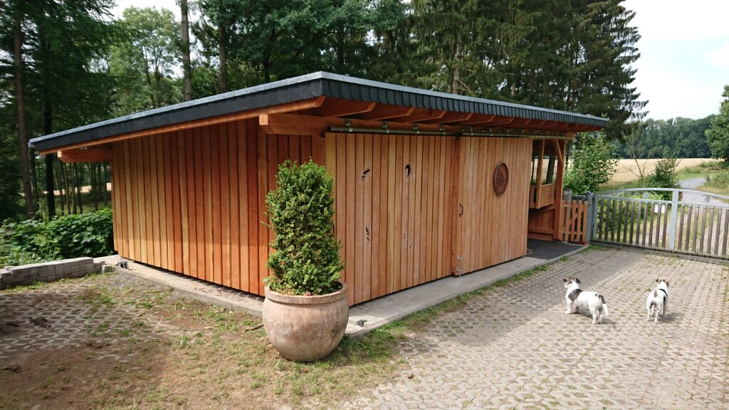 Hölzerne Gartenhütte mit zwei Hunden davor, umgeben von Wald und Feld in ländlicher Umgebung.