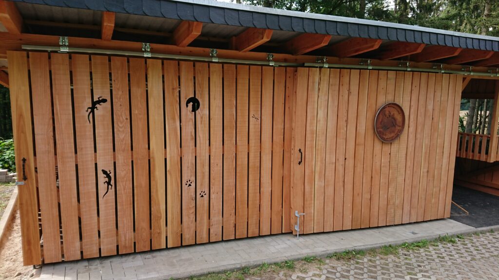 Holzwand mit Tiermotiven und rundem Schild, dunkles Schindeldach, gepflasterter Boden.