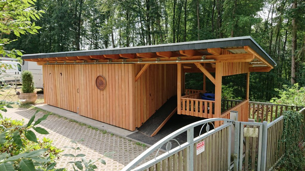 Ein hölzerner Schuppen mit Metallzaun, umgeben von viel Grün, steht in einer ruhigen, ländlichen Umgebung.