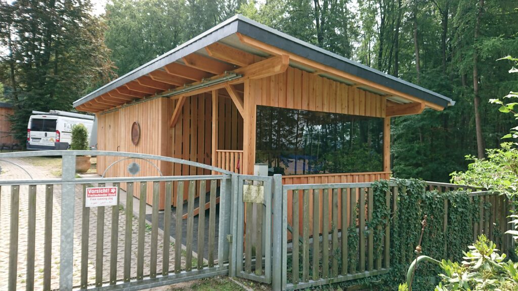 Moderne Holzhütte im Wald mit großem Fenster, Metallzaun und Schild „Vorsicht! Freilaufender Hund“.