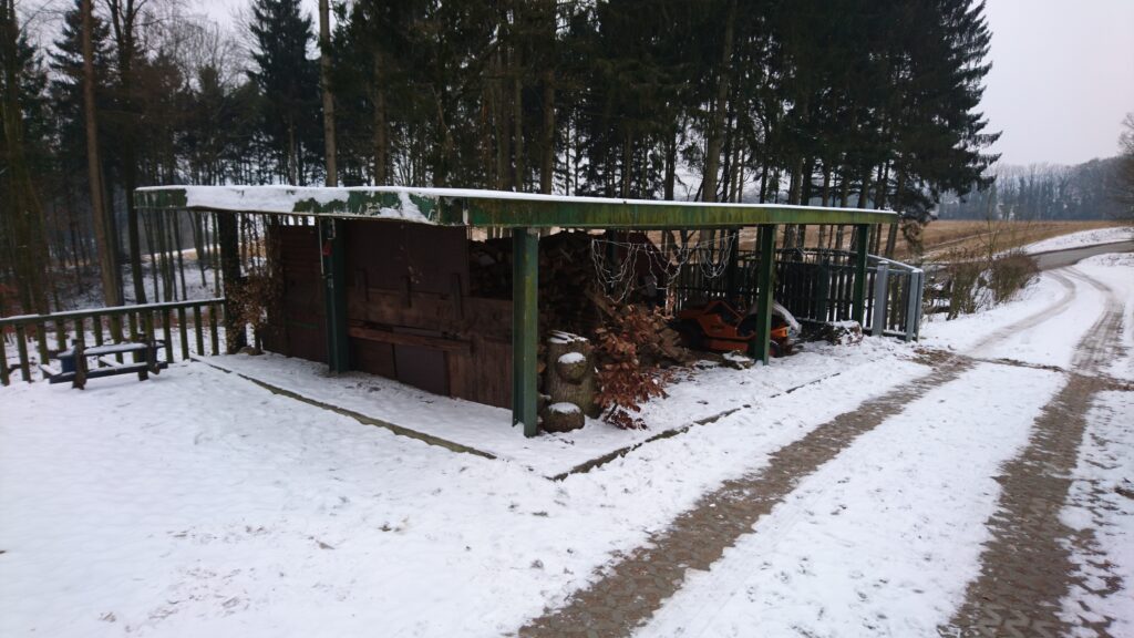 Winterlicher Holzunterstand mit Brennholz und orangefarbenem Gerät, umgeben von Schnee und Bäumen.