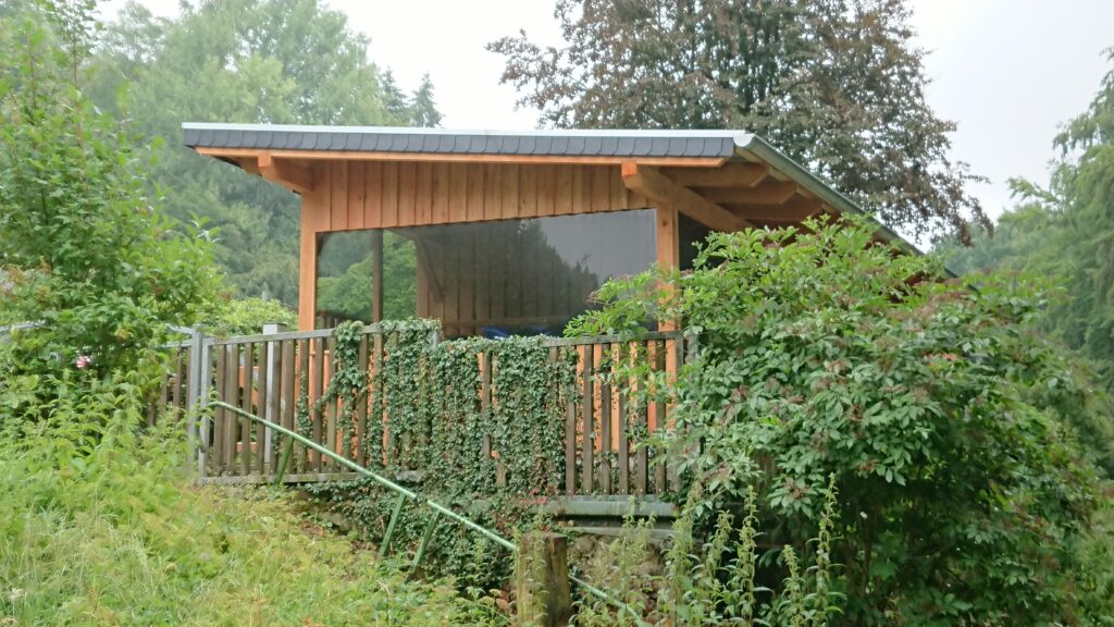 Ein kleines Holzgebäude steht inmitten grüner Vegetation, umgeben von einem bewachsenen Zaun.
