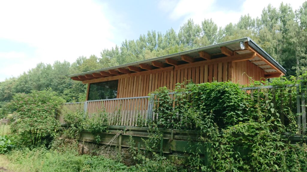Modernes Holzgebäude in grüner Umgebung, umgeben von üppiger Vegetation und blauem Himmel.