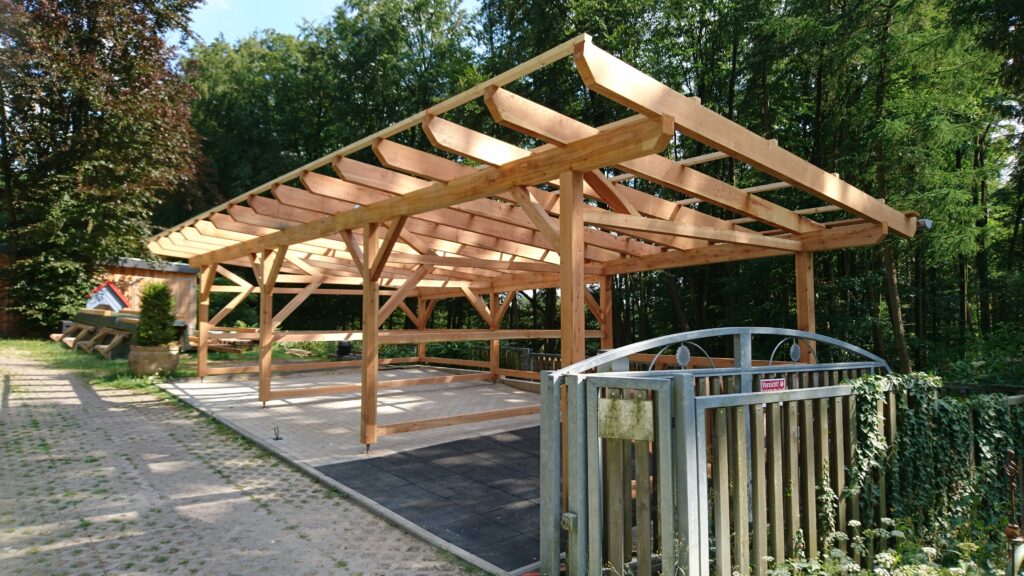 Teilweise gebaute Holzkonstruktion in natürlicher Umgebung, umgeben von Bäumen und einem Metallzaun.