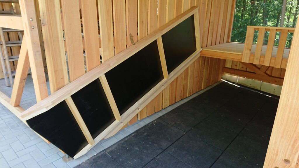 Holzklettergerüst mit schräger Kletterwand und Plattform, umgeben von grauem Bodenbelag.