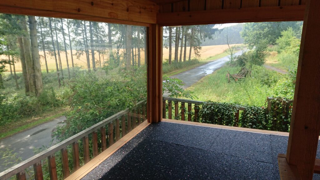 Überdachte Holzveranda mit Blick auf grüne Landschaft und bewölkten Himmel, friedliche Atmosphäre.