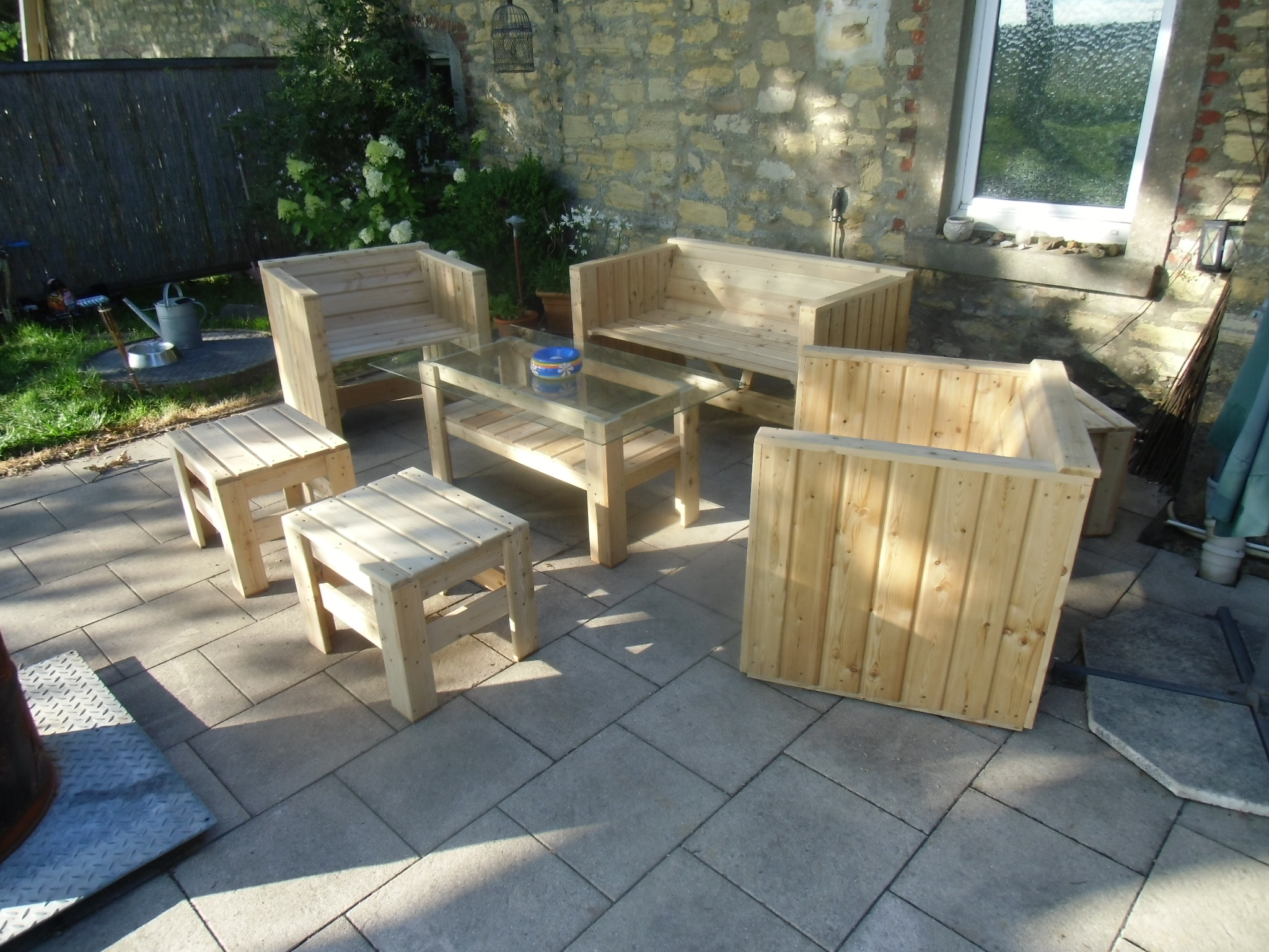 Terrasse mit Holzmöbeln, darunter Bänke und ein Tisch, umgeben von Pflanzen und einer Steinwand.