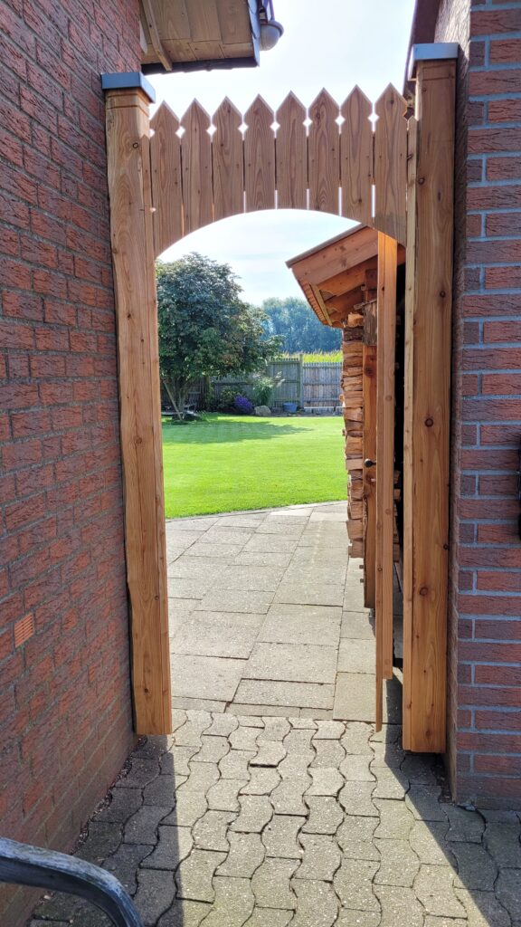 Ein hölzernes Tor zwischen Backsteinmauern führt zu einem gepflegten Garten mit grünem Rasen.
