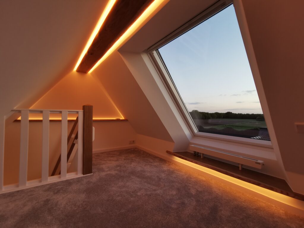 Moderner Dachboden mit großem Fenster, warmem Licht und Blick auf Sonnenuntergang über Bäumen.