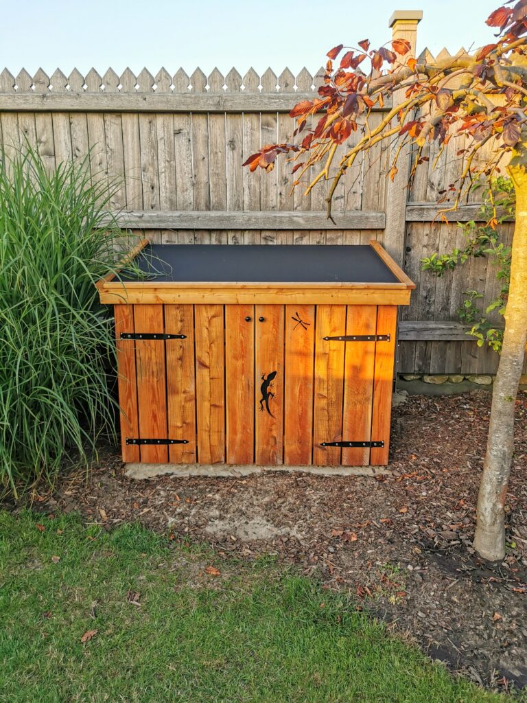 Ein Holzkasten im Garten, umgeben von einem Holzzaun, einem Baum mit rötlichen Blättern und grünem Gras.