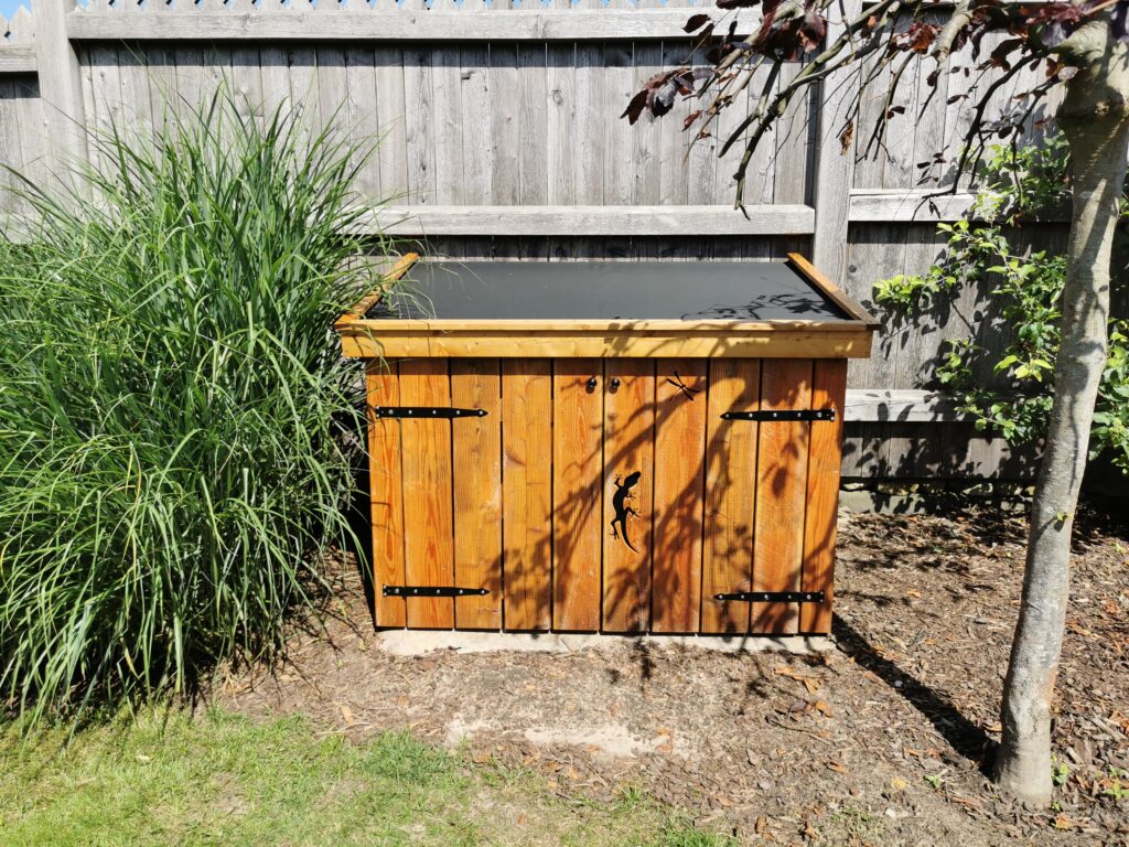 Ein hölzerner Schuppen mit Geckosymbol steht in einem grünen Garten neben einem grauen Holzzaun.