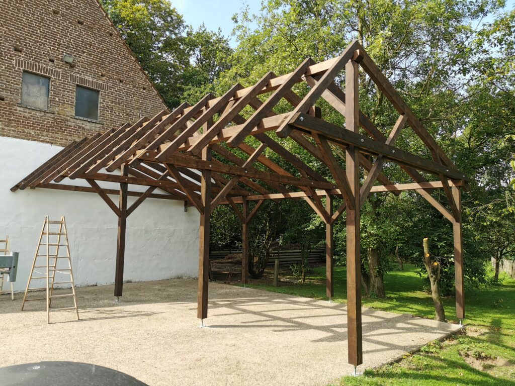 Holzgerüst im Bau, umgeben von Bäumen und einem gemauerten Gebäude, mit Leiter daneben.