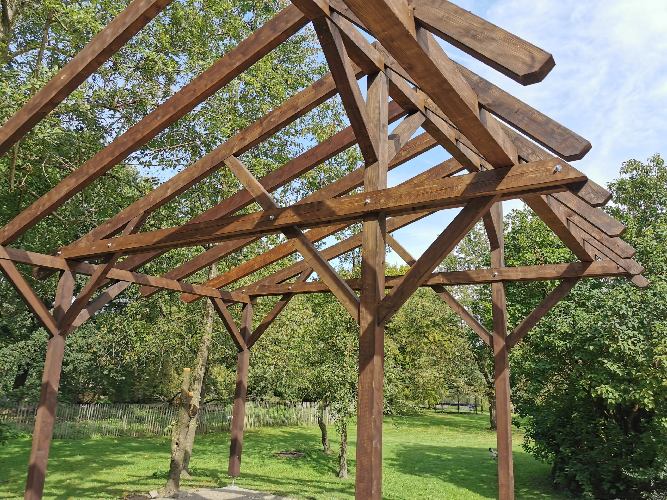 Holzgerüst eines Pavillons vor grüner Wiese und Bäumen, unter teils bewölktem Himmel.
