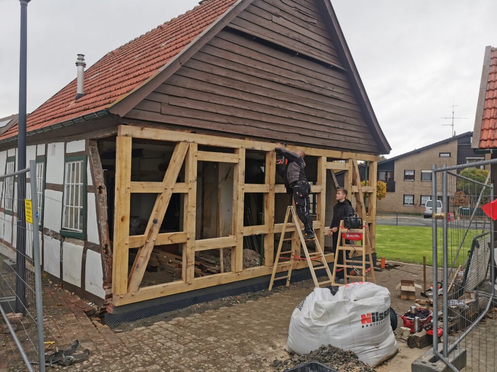 Fachwerkhaus-Renovierung: Zwei Arbeiter auf Baustelle, sichtbares Holzgerüst, bewölkter Himmel.