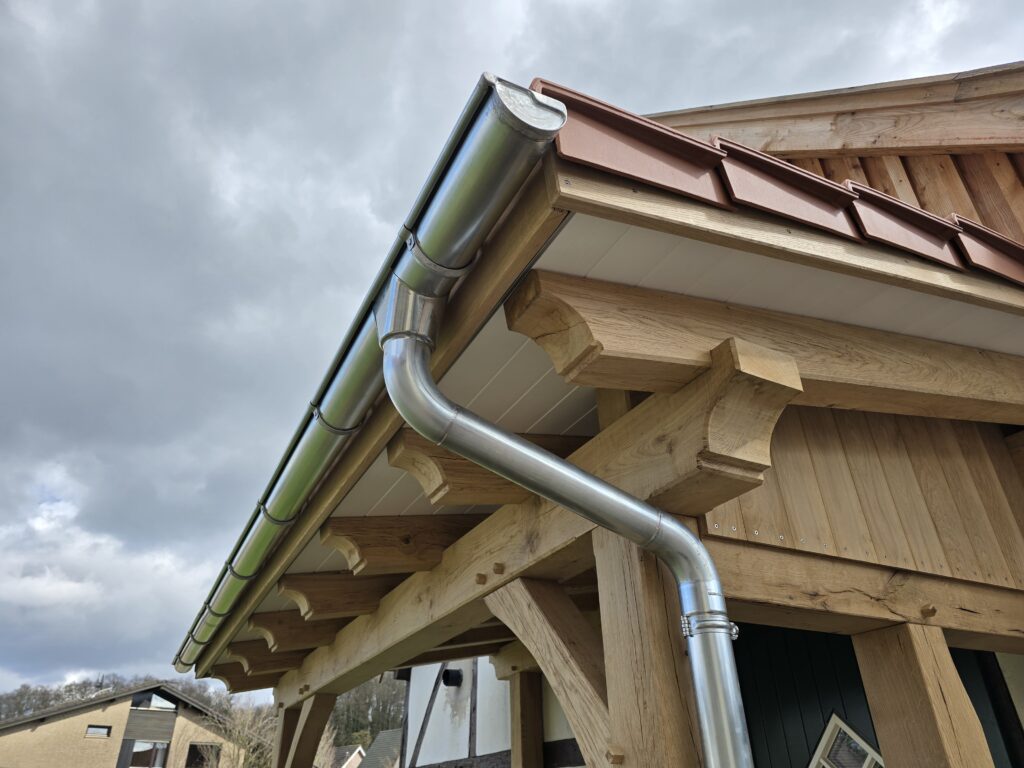 Ein modernes Dach mit roten Ziegeln, silberner Dachrinne und massiver Holzstruktur bei bewölktem Himmel.