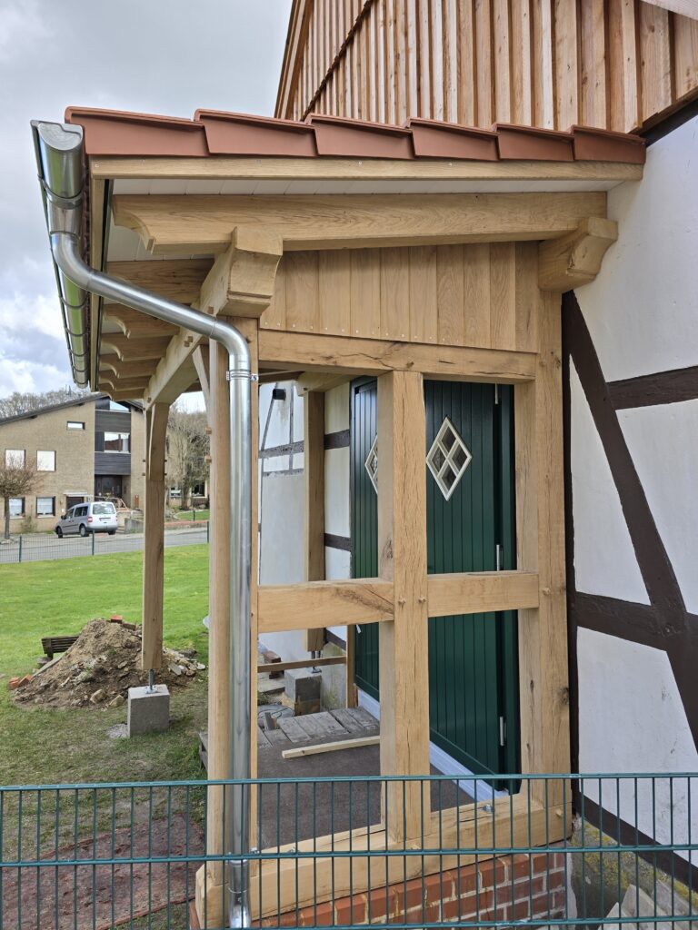 Eingangsbereich eines Fachwerkhauses mit grünem Tür, Holzvordach und sichtbaren Bauarbeiten.