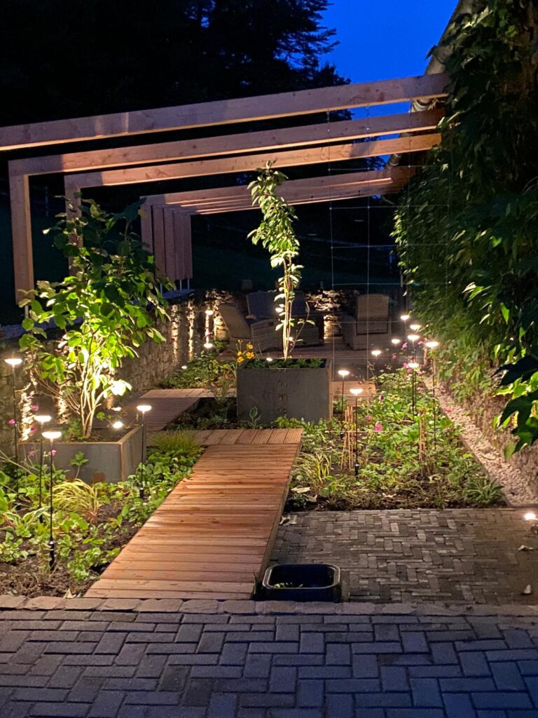 Ein stilvoll beleuchteter Garten mit Holzweg, Pergola und Pflanzkästen in ruhiger Abendstimmung.