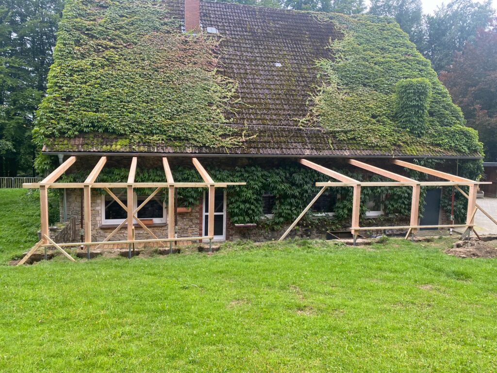 Ein Haus mit efeubewachsenem Dach und einer im Bau befindlichen Holzkonstruktion im Vordergrund.