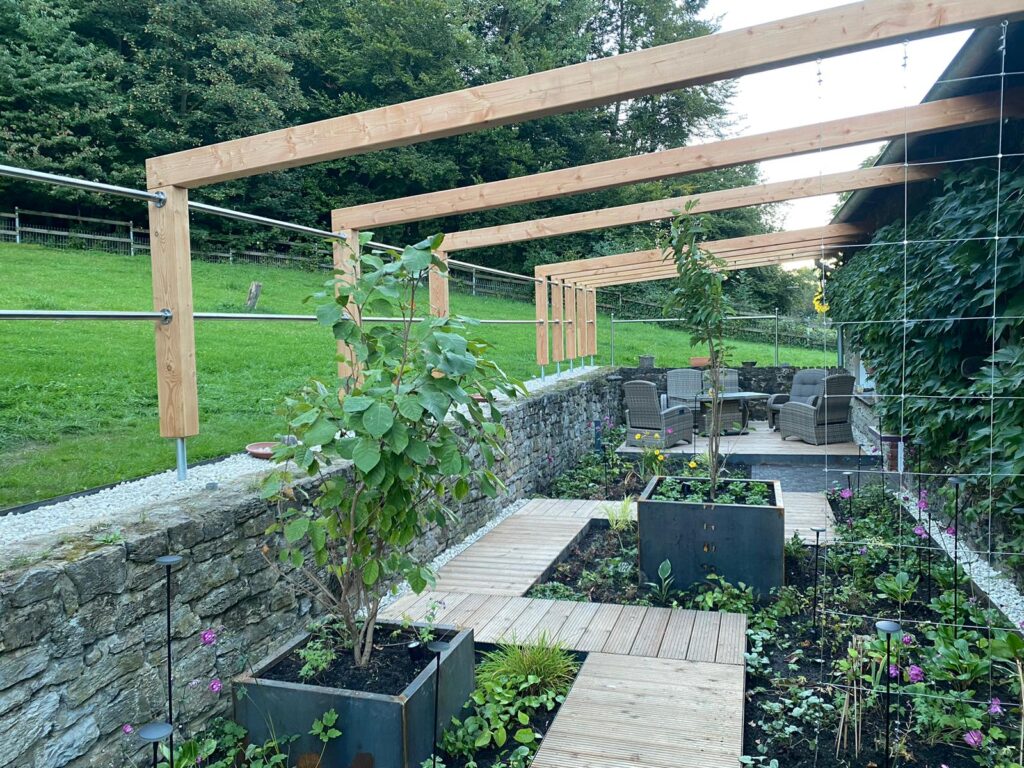 Ein gepflegter Garten mit Holzpergola, Steinmauer, Pflanzkübeln und Rattan-Gartenmöbeln auf Terrasse.