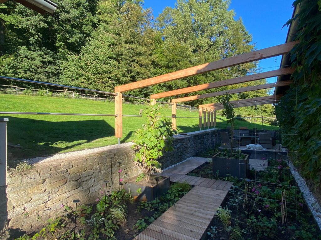 Ein gepflegter Garten mit Pergola, Hochbeeten, Sitzbereich und einem Hund, der entspannt liegt.