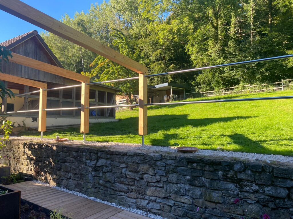 Ländliche Szene mit Holzhaus, modernem Geländer, Steinmauer, grüner Wiese und Wald im Hintergrund.