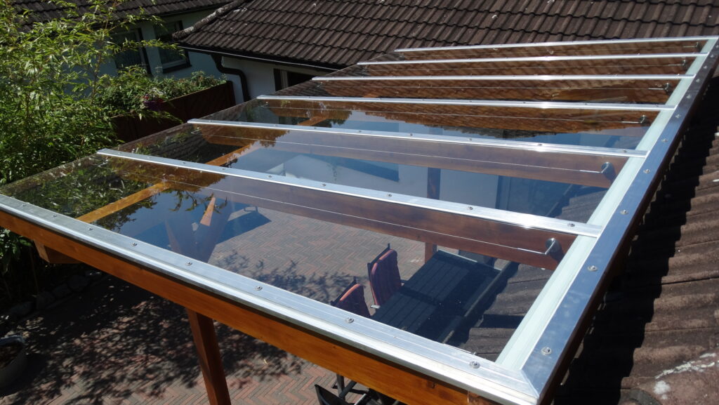 Terrassenüberdachung aus Glas mit Holz- und Metallstreben, darunter Tisch und Stühle auf Pflastersteinen.