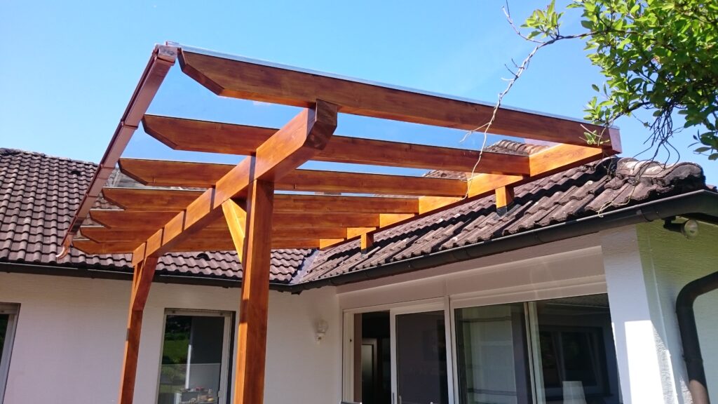 Eine moderne Holzpergola ergänzt ein Haus mit dunklem Ziegeldach und weißer Fassade unter blauem Himmel.