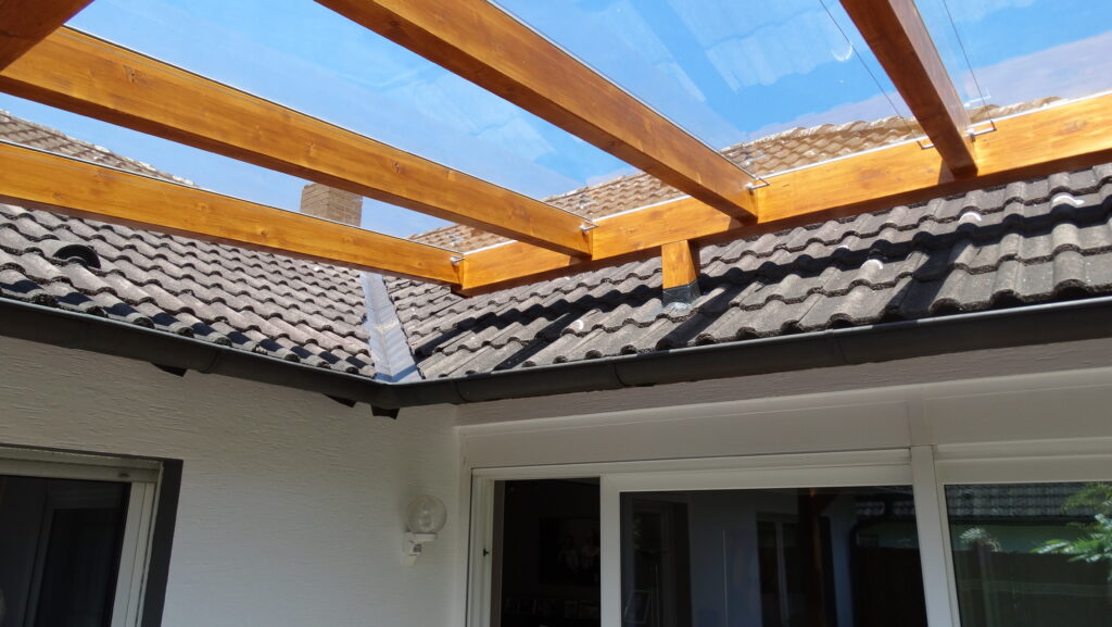 Überdachte Terrasse mit Holzbalken und Glasdach, angrenzend an ein Haus mit dunklen Dachziegeln.