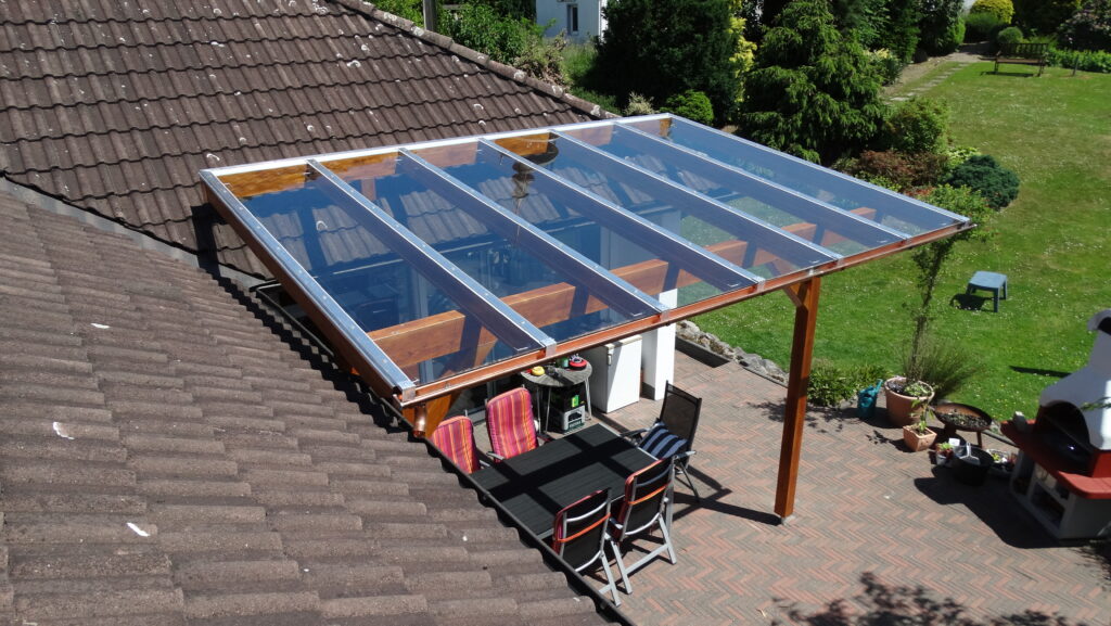 Überdachte Terrasse mit Tisch, bunten Kissen, gemauertem Grill und gepflegtem Garten im Sonnenschein.
